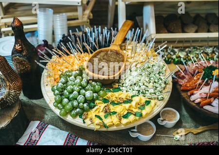 Plateau rond en bois avec des morceaux de différents types de fromage sur brochettes et sauce au milieu. Banque D'Images