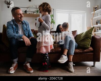 Papa et fille souriant pendant qu'elle montre ses devoirs tandis que maman et sœur s'assoient confortablement sur le canapé du salon. Banque D'Images