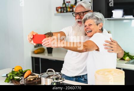 Couple senior heureux prenant le selfie tout en cuisinant ensemble à la maison - les personnes âgées s'amusent à préparer un déjeuner santé dans la cuisine Banque D'Images