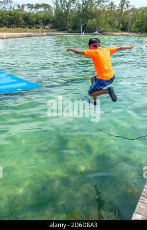 Garçon portant un t-shirt orange et un short de bain sautant d'un quai dans les eaux turquoise des Florida Keys à Key Largo lors d'une journée ensoleillée. Banque D'Images