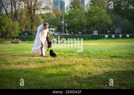 Soin de la barbe de grande qualité, seric portant une garbe sacerdotale blanche traversant le parc de l'église vers le chat errant après le service divin. Banque D'Images