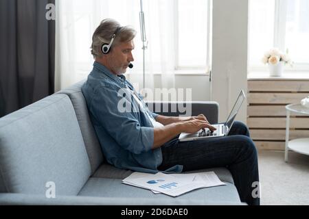 Homme positif dans les écouteurs utilisant un ordinateur portable écouter un podcast ou participer à un webinaire assis sur un canapé à la maison. Banque D'Images