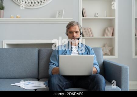 Homme positif dans les écouteurs utilisant un ordinateur portable écouter un podcast ou participer à un webinaire assis sur un canapé à la maison. Banque D'Images