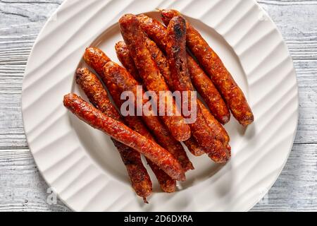 Petites saucisses françaises chipolata grillées et servies sur une assiette blanche sur fond de bois blanc, vue de dessus, gros plan Banque D'Images