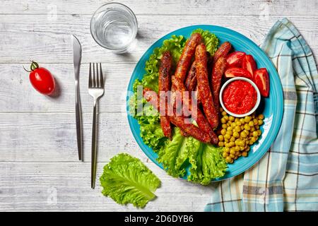 Petites saucisses de porc italiennes Chipolata grillées et servies sur une assiette bleue avec ketchup de tomate et tomates cerises, laitue verte et petits pois sur une W blanche Banque D'Images