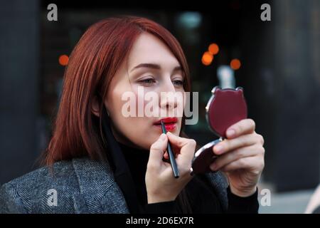 Jolie femme à tête rouge regarde le miroir de poche et applique rouge à lèvres avant la réunion d'affaires. Notion datation après travail. Banque D'Images