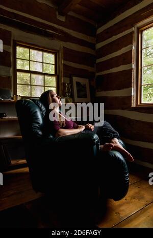 Femme se reposant dans un fauteuil inclinable en cuir dans une maison rustique en bois rond. Banque D'Images