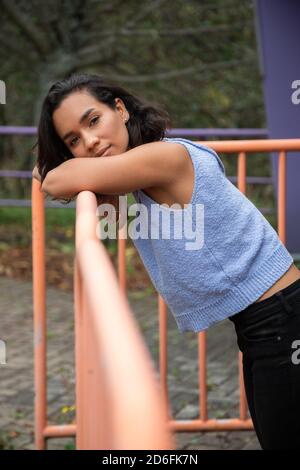 Jeune femme brunette Latina avec des cheveux courts et des boucles, s'appuyant contre la main courante, portant un t-shirt bleu et un pantalon noir, lumière naturelle et décontractée Banque D'Images