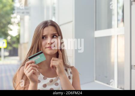 Jolie jeune femme avec carte de crédit touchant la joue et regardant loin en se tenant sur la rue de la ville et de penser Banque D'Images