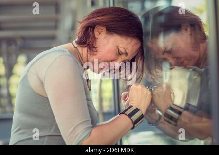 Vue latérale d'une femme sénior attirante qui garde les yeux fermés et penchée sur un mur de verre avec une expression triste du visage Banque D'Images