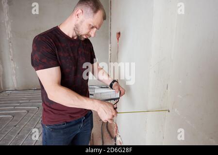 Gros plan d'un jeune professionnel dans un appartement vide inachevé mesurant le mur à l'aide d'un ruban de mesure, en vérifiant les notes. Concept de réparation et de remise à neuf Banque D'Images