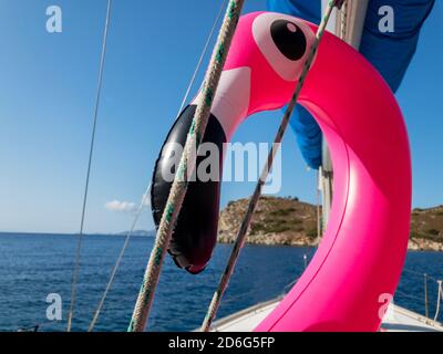 Rose flamant plastique gonflable avec expression drôle sur un bateau à voile. Fond bleu de mer et de ciel Banque D'Images