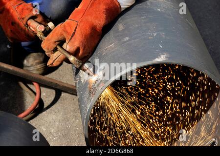 Le soudeur soudeur à la main soudeur un tube de tuyau métallique large avec un chalumeau de découpe à l'oxy-fuel, avec flamme et étincelles, gros plan sur le chantier de construction Banque D'Images