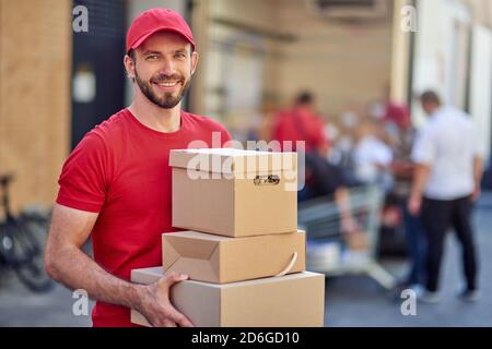 Jeune homme caucasien souriant à la caméra tout en tenant des boîtes en papier pour la livraison debout à la maison de stockage. Concept de livraison de nourriture Banque D'Images