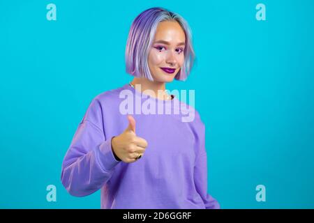 Femme mignonne avec une coiffure teinte violette montrant le signe de pouce vers le haut sur fond bleu. Une jeune fille positive sourit à l'appareil photo. Gagnant. Réussite. Langage corporel Banque D'Images