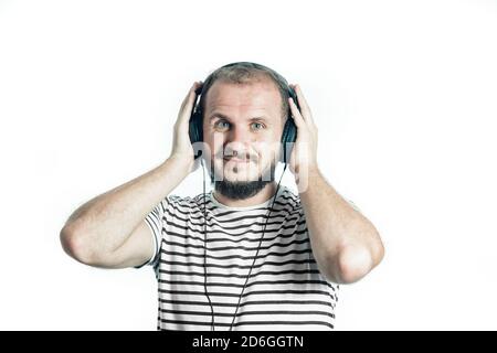 Souriant barbu heureux homme dans un t-shirt rayé écoute de la musique avec de grands écouteurs. Regardez la caméra. Isolé sur blanc. Banque D'Images