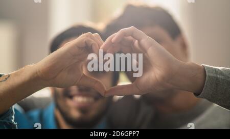 Portrait d'un couple de Quéer hommes mignon à la maison. Ils s'assoient sur un canapé et font un symbole de coeur avec leurs mains. Le partenaire a une main derrière son Lover Banque D'Images