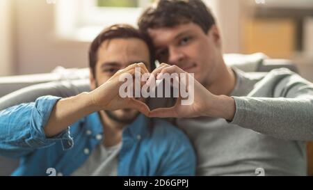 Portrait d'un couple de Quéer hommes mignon à la maison. Ils s'assoient sur un canapé et font un symbole de coeur avec leurs mains. Le partenaire a une main derrière son Lover Banque D'Images