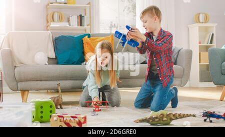 Dans le salon: Garçon et fille jouant avec les avions et les dinosaures tout en étant assis sur un tapis. Salon ensoleillé avec enfants s'amuser. Banque D'Images