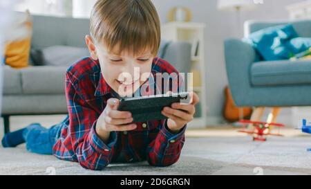 Smart Little Boy pose sur un tapis joue dans le jeu vidéo sur son smartphone, tient le téléphone mobile en mode horizontal Paysage. L'enfant a du plaisir à jouer Banque D'Images