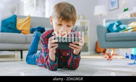 Smart Little Boy pose sur un tapis joue dans le jeu vidéo sur son smartphone, tient le téléphone mobile en mode horizontal Paysage. L'enfant a du plaisir à jouer Banque D'Images