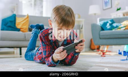 Smart Little Boy pose sur un tapis joue dans le jeu vidéo sur son smartphone, tient le téléphone mobile en mode horizontal Paysage. L'enfant a du plaisir à jouer Banque D'Images