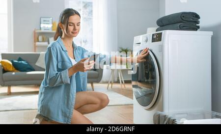 Belle jeune femme est assise à genoux à côté de la machine à laver. Elle a chargé le lave-linge avec du linge sale tout en utilisant son smartphone. Tourné vers l'intérieur Banque D'Images