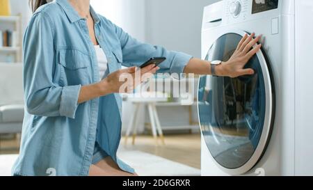 Belle jeune femme est assise à genoux à côté de la machine à laver. Elle a chargé le lave-linge avec du linge sale et a configuré le lave-linge avec elle Banque D'Images