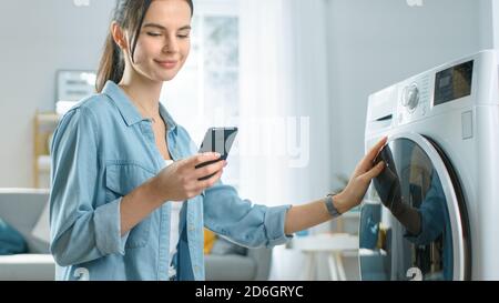 Belle jeune femme est assise à genoux à côté de la machine à laver. Elle a chargé le lave-linge avec du linge sale et a configuré le lave-linge avec elle Banque D'Images