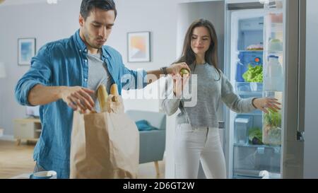 Un jeune couple magnifique vient à la cuisine avec des provisions fraîches dans un sac de papier brun. L'homme donne des pommes et des oranges fraîches à la fille qui les met Banque D'Images