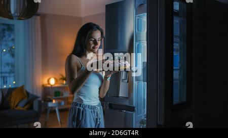 Une jeune femme affamée dans la cuisine le soir a ouvert le réfrigérateur, prend un morceau d'un délicieux gâteau crémeux et commence à l'est Banque D'Images