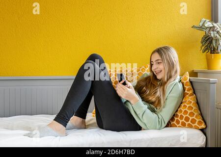 Jeune fille adolescente blonde utilisant un smartphone sur le lit à l'avant d'un mur jaune Banque D'Images