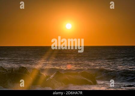 Vue idyllique des vagues se brisant contre les rochers au coucher du soleil contre le ciel orange et coucher du soleil avec les rayons du soleil et la tache solaire Banque D'Images