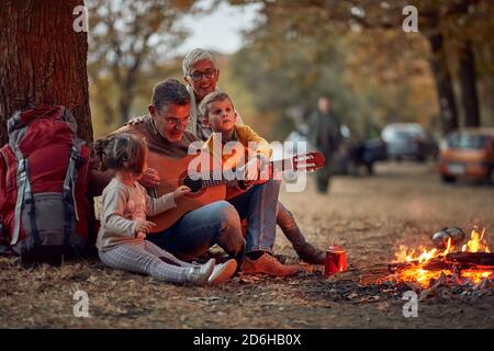 Un couple âgé et des petits-enfants jouant de la guitare dans la forêt autour d'un feu de camp sur un beau crépuscule d'automne Banque D'Images