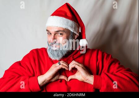 le père noël dans le sweat à capuche forme un cœur avec son mains devant sa poitrine et regarde le appareil photo avec un sourire amical Banque D'Images