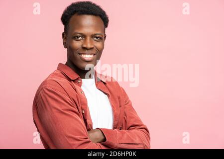 Souriant assuré Noir homme en chemise rouge debout avec bras croisés, regardant l'appareil photo avec bonne humeur, sur fond rose studio Banque D'Images