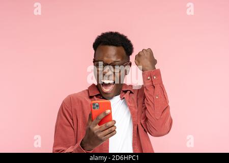 Un homme millénaire afro-américain excité en lunettes tient le téléphone portable isolé sur fond rose de studio. Un homme noir joyeux regarde le smartphone, sourit fe Banque D'Images
