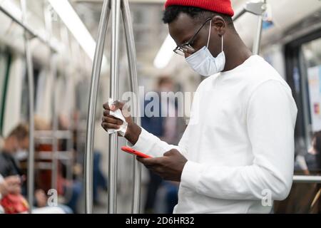 Homme millénaire noir en chapeau rouge portant un masque de protection contre le covid-19, virus de la grippe, en utilisant un téléphone mobile, tenant une main courante dans les transports publics Banque D'Images