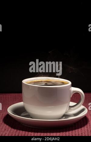 Café cuisant dans une tasse avec soucoupe isolée sur un arrière-plan noir sur une surface rouge Banque D'Images