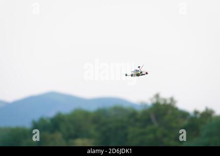 Les hélices colorées soulèvent ce drone de cotter FPV de course à travers les airs. Fond ciel blanc Uni avec des collines et des arbres flous en dessous Banque D'Images