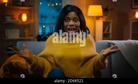 Portrait d'une belle fille noire assise sur un canapé à la maison la nuit, regardant un film à la télévision. Elle est vraiment excitée et émotive. Banque D'Images