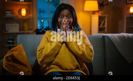 Portrait d'une belle fille noire assise sur un canapé à la maison la nuit, regardant un film d'horreur à la télévision. Elle est vraiment effrayée et émotive. Banque D'Images