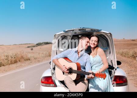 Joyeux jeune couple jouant de la guitare et de profiter du temps ensemble assis sur le coffre ouvert de la voiture garée sur la route de campagne en été Banque D'Images