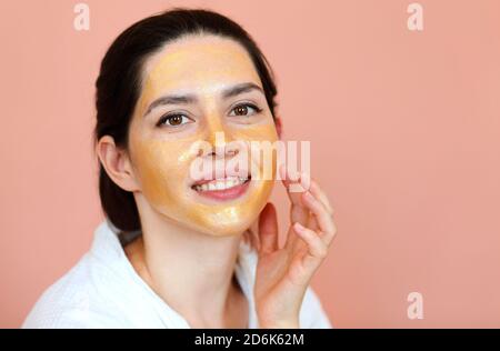 Charmant contenu femme en peignoir blanc et avec masque facial regarder la caméra tout en faisant la procédure de beauté sur fond rose Banque D'Images