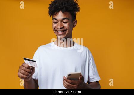 Image de joyeux afro-américain gars tenant la carte de crédit et téléphone portable isolé sur un mur jaune Banque D'Images