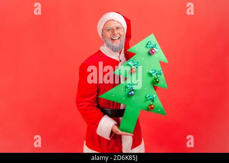 Joyeux Père noël âgé avec une barbe grise en lunettes tenant un arbre de noël décoré de papier, préparant les vacances d'hiver. Studio d'intérieur tourné isol Banque D'Images