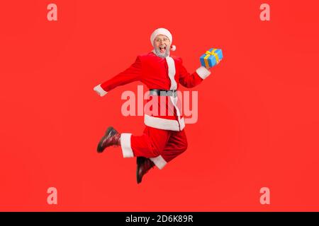 Le Père noël est extrêmement heureux et tient une boîte cadeau sautant et regardant l'appareil photo avec un sourire éclatant, satisfait des vacances d'hiver. Prise de vue en studio Banque D'Images