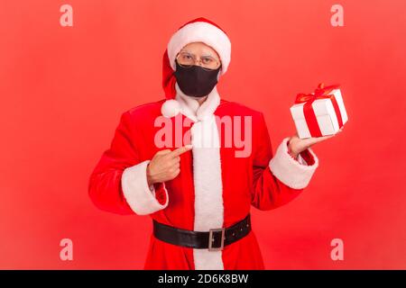 Homme âgé en costume de fête du père noël portant un masque facial protecteur pointant le doigt sur la boîte à cadeaux dans sa main, préparant des cadeaux pendant les vacances. IND Banque D'Images