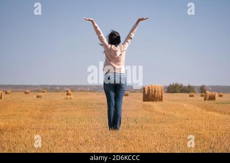 Une fille heureuse, libre, indépendante, rêveuse sur le terrain a soulevé ses mains au ciel. Une femme dans une veste et un Jean à la vue arrière de la moisson. Silhouette o Banque D'Images