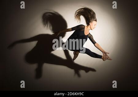 la femme dans l'obscurité effectue le saut de gymnastique, sur un fond blanc il y a une ombre d'une forme. Photographie en studio. Banque D'Images